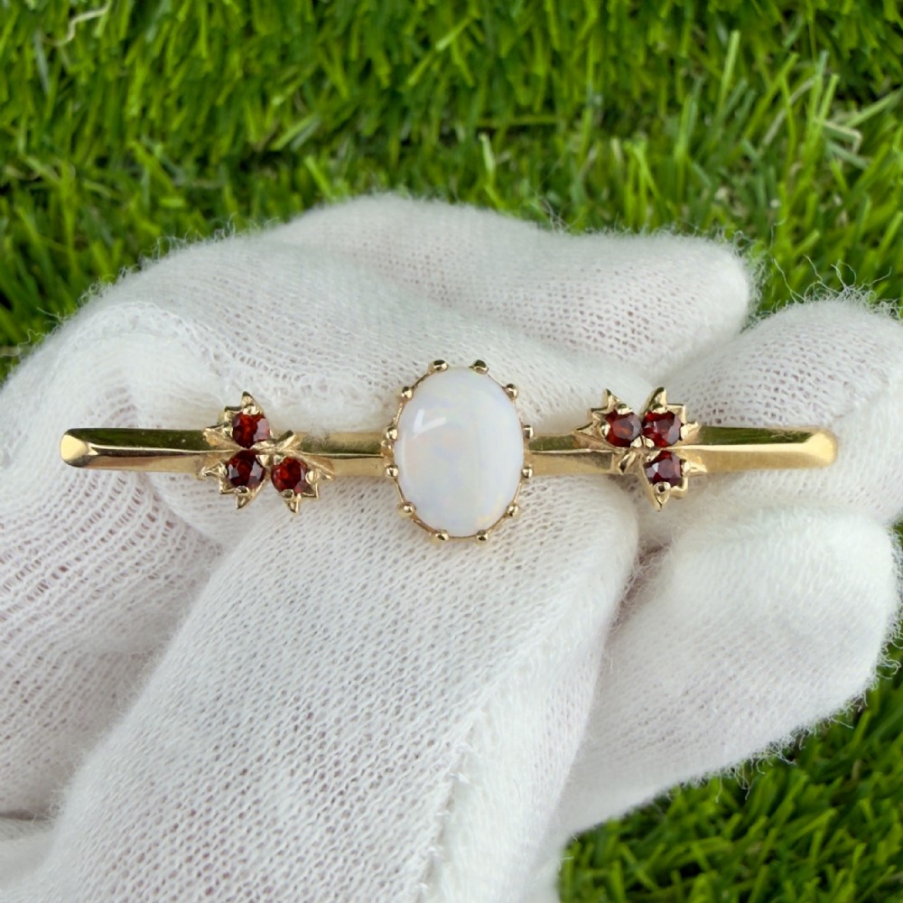 A yellow gold bar brooch featuring a central oval cabochon opal flanked by two cluster arrangements of red garnets.