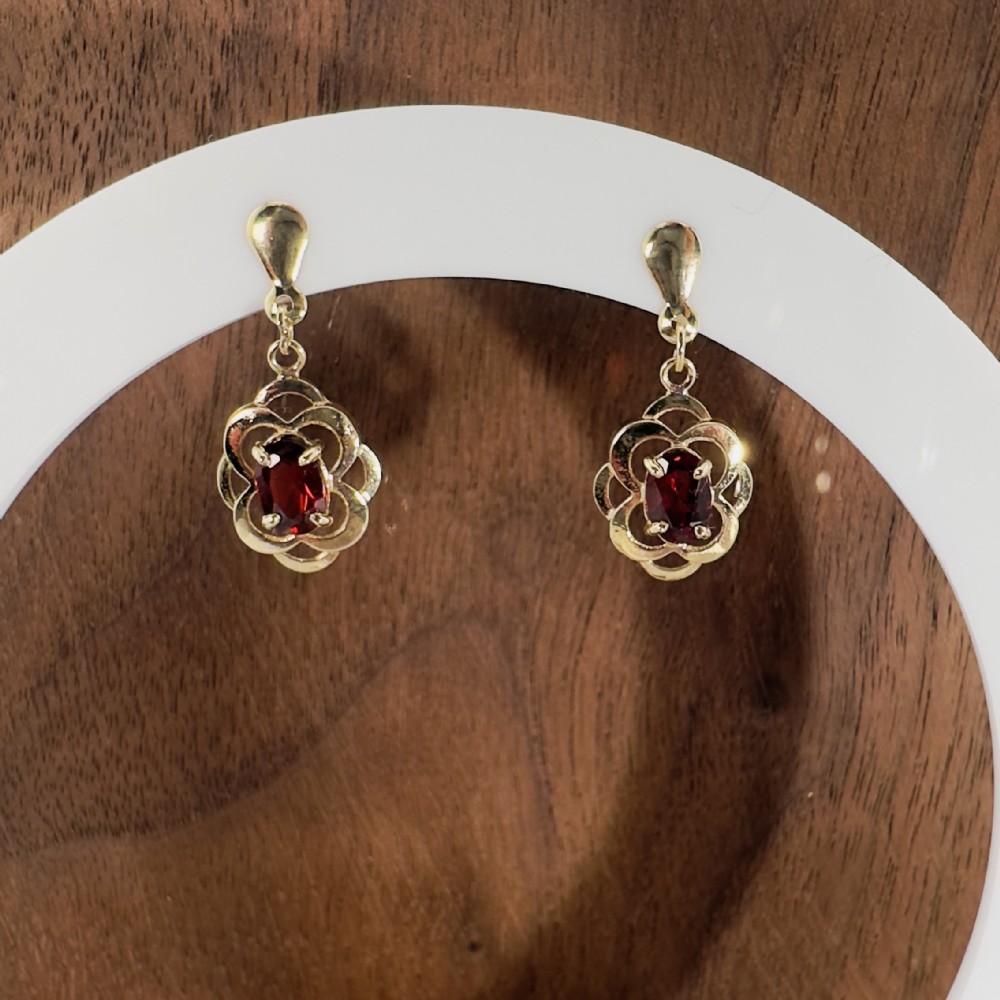 A pair of 9ct yellow gold drop stud earrings featuring oval red garnets framed by openwork floral metalwork.