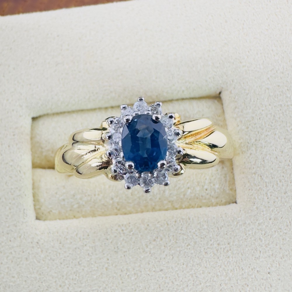 An 18ct yellow gold ring featuring a central oval blue sapphire surrounded by a halo of small diamonds and set on fluted shoulders.