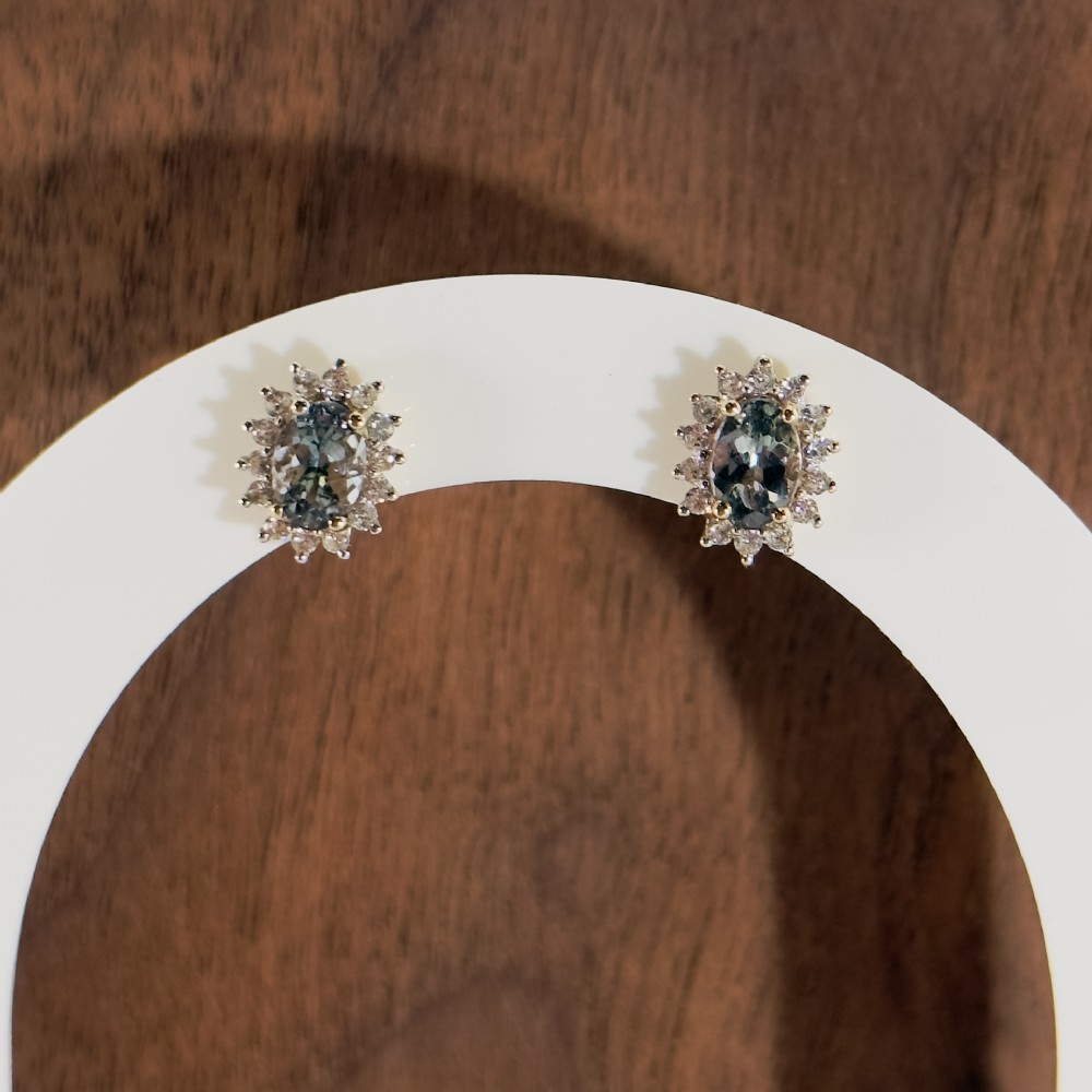 A pair of 9ct yellow gold stud earrings featuring central oval cut andalusite gemstones surrounded by a halo of small diamonds.