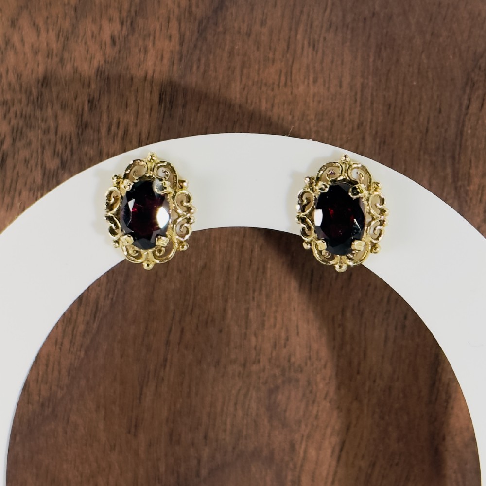 A pair of 9ct yellow gold stud earrings featuring central oval-cut deep red garnets encased in ornate filigree borders.