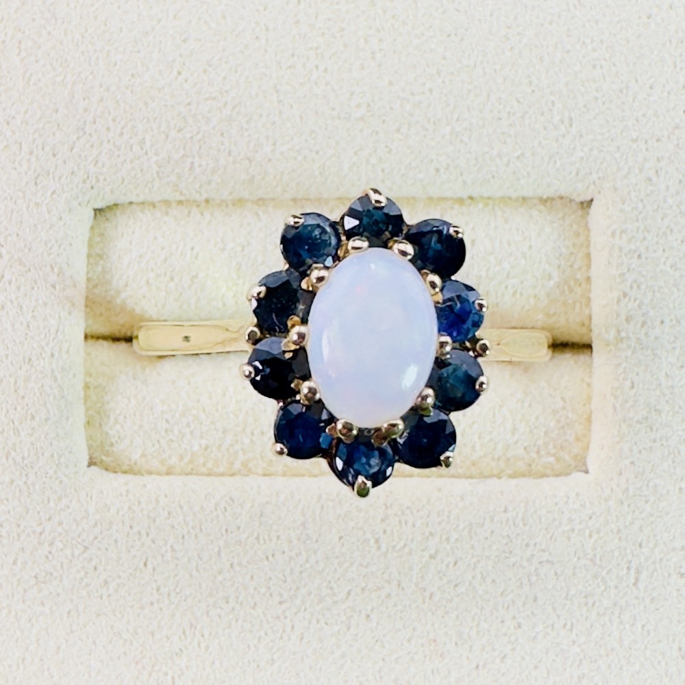 A 9ct yellow gold ring featuring a central oval cabochon opal surrounded by ten round blue sapphires in claw settings.