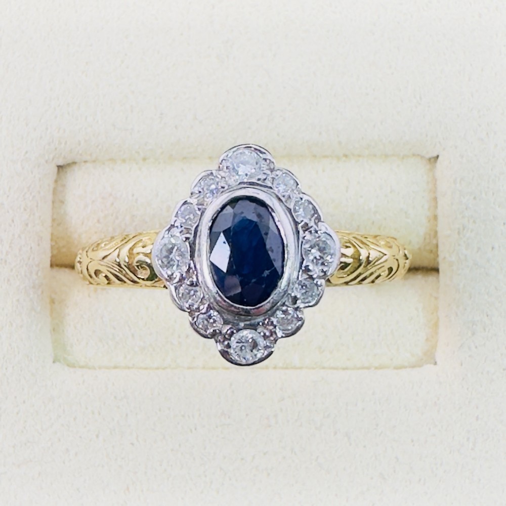 An 18ct yellow gold ring featuring an oval cut deep blue sapphire framed by a halo of diamonds on an intricately engraved band.