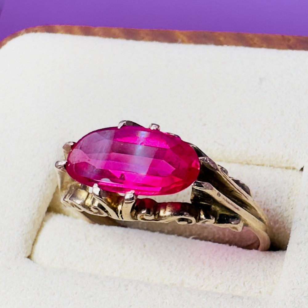 A 14ct gold ring featuring an elongated oval-cut pinkish-red ruby held in a multi-claw setting with decorative scrollwork on the side gallery.