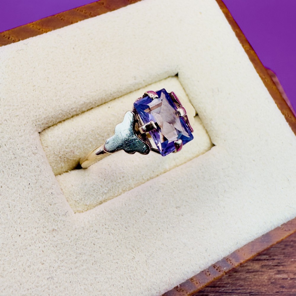 A 9ct gold Art Deco solitaire ring featuring a rectangular emerald-cut purple sapphire set with four double claw corner mounts and stepped shoulder details.