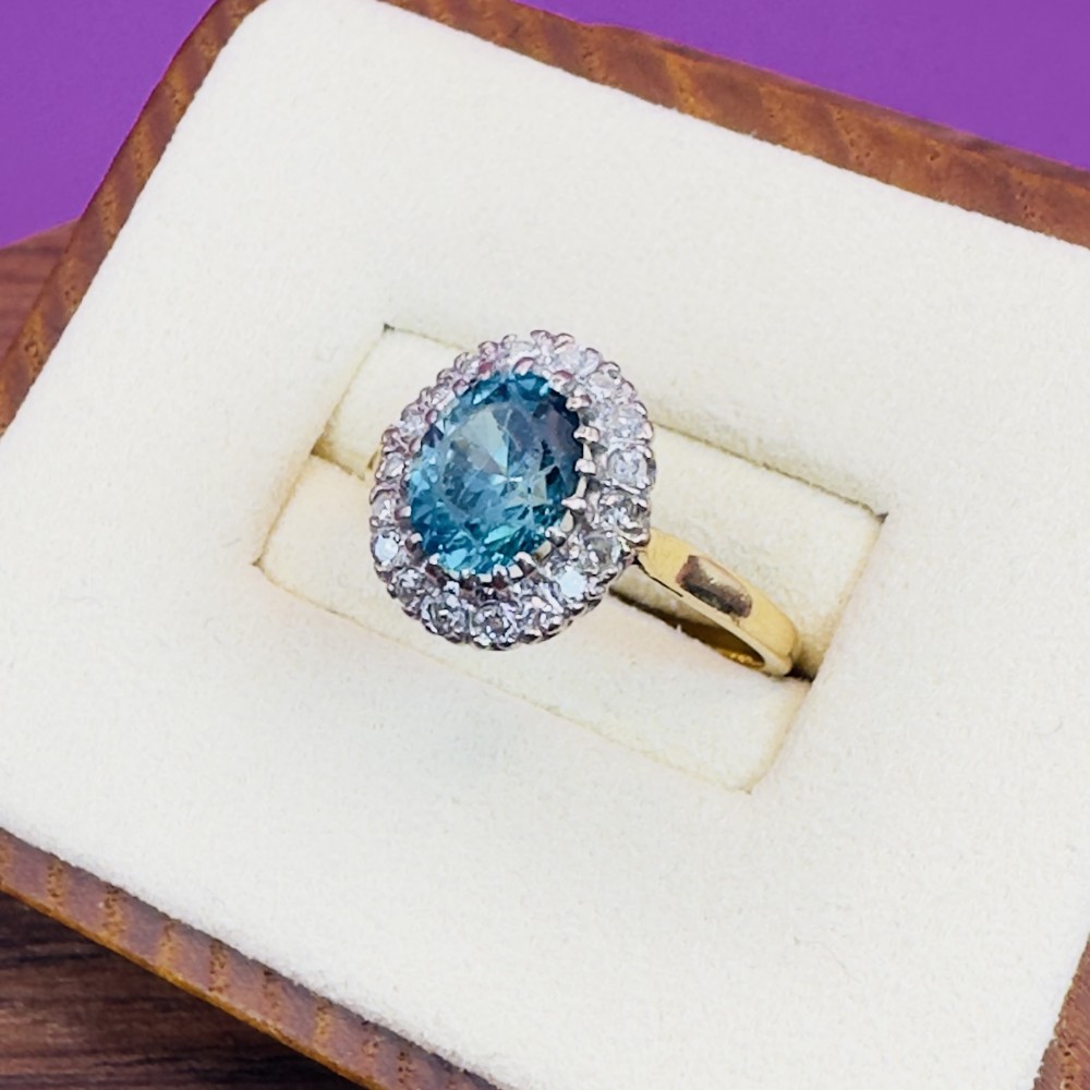 An Art Deco cluster ring featuring a central oval blue zircon gemstone surrounded by a halo of round diamonds set on an 18ct yellow gold band.