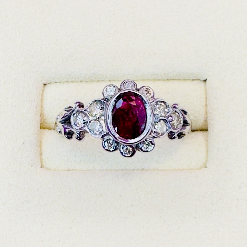 An 18ct white gold cluster ring featuring a central oval red ruby surrounded by small round diamonds and carved shoulder details.