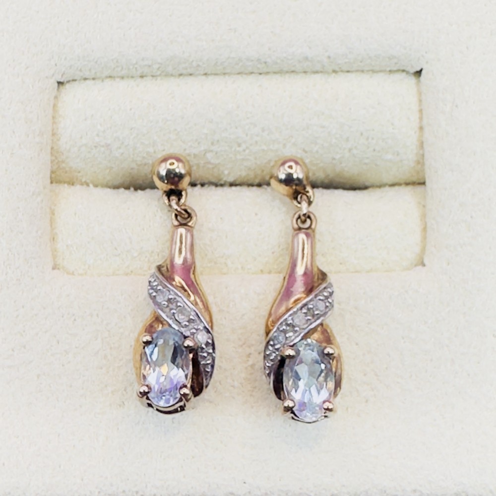 Pair of 9ct yellow gold drop stud earrings featuring oval-cut light blue aquamarines enclosed in diamond-paved swirl mounts.