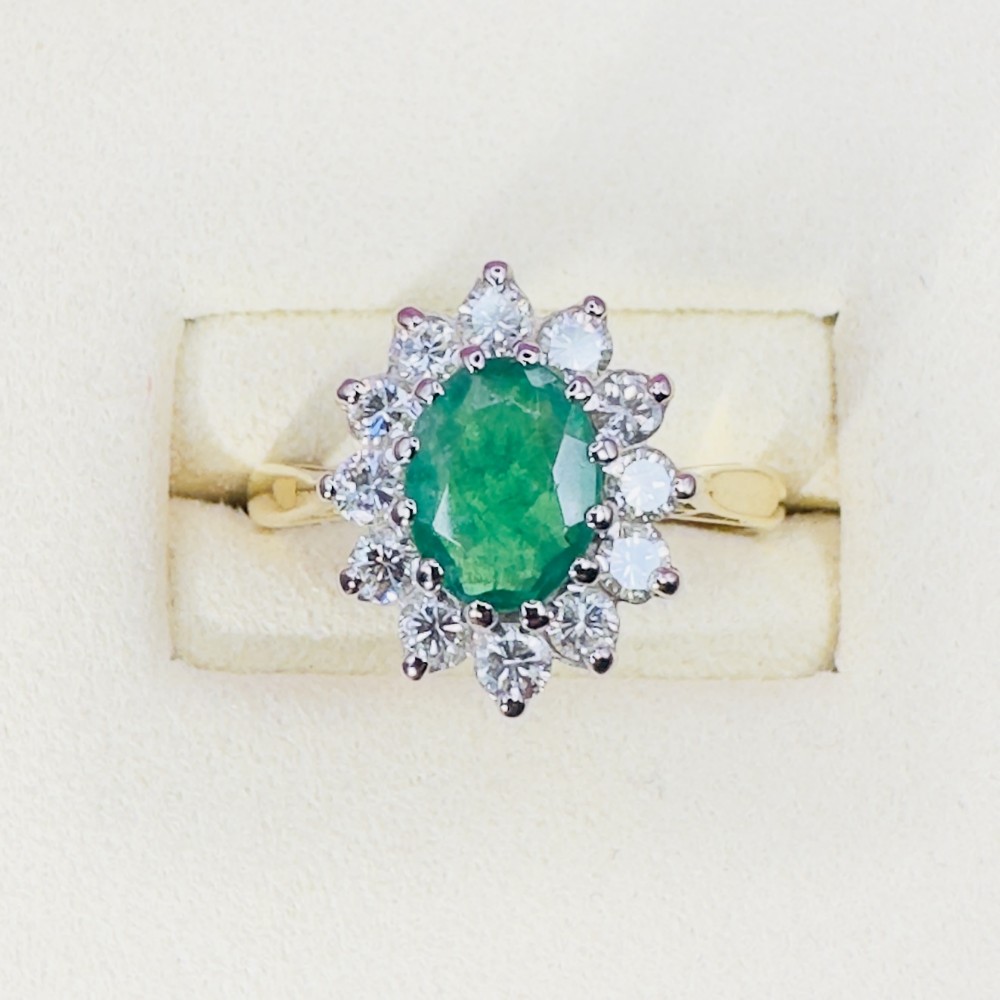 An 18ct yellow gold ring set with an oval faceted central green emerald and a halo cluster of round brilliant diamonds.
