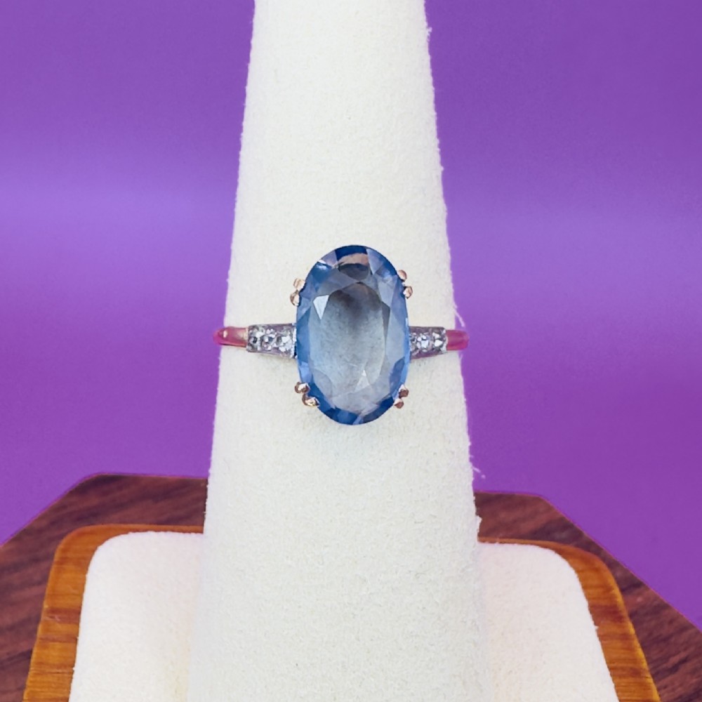 An Art Deco ring featuring a large oval cut blue sapphire in claw settings with diamond shoulders on a gold band.
