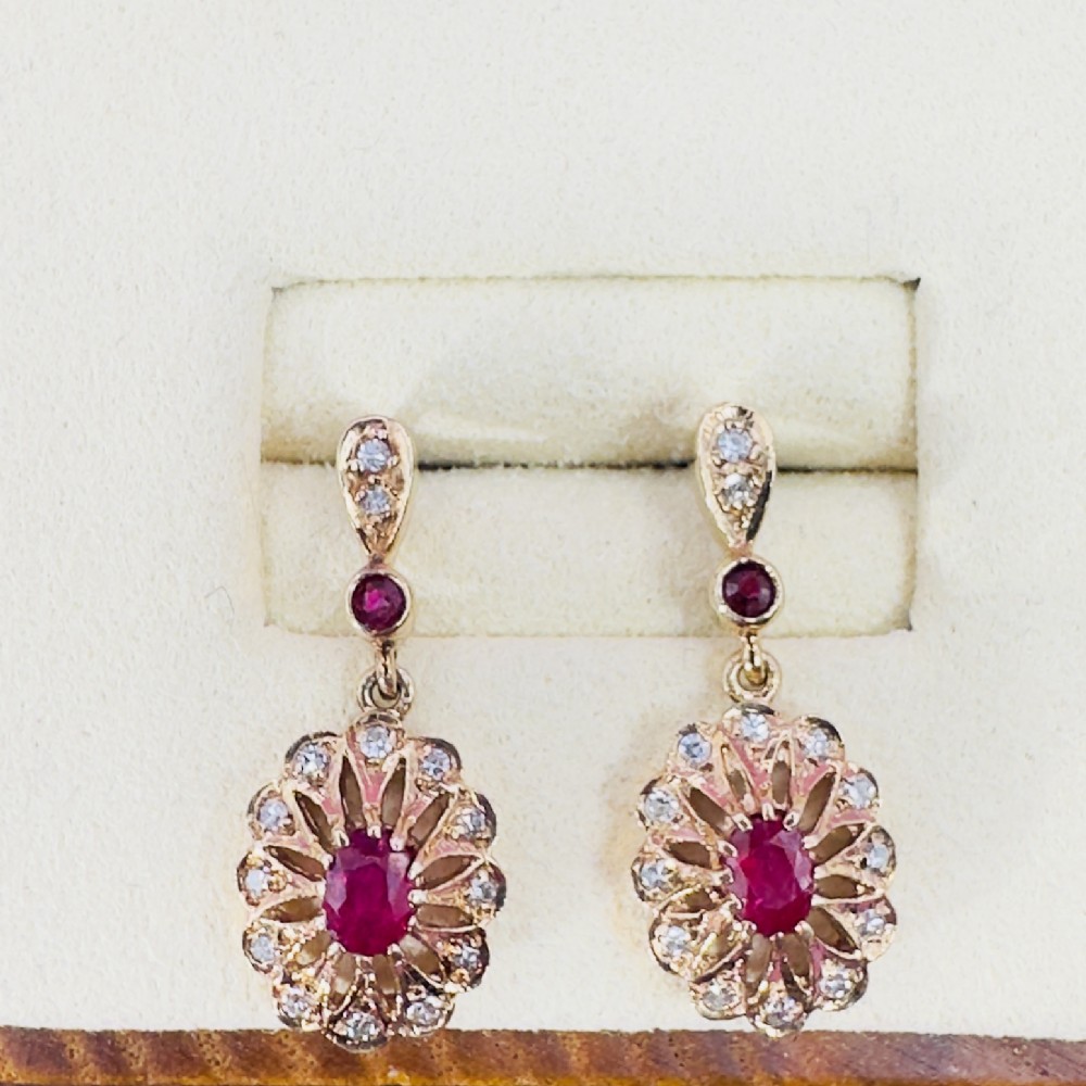 A pair of 9ct gold drop stud earrings featuring central oval red rubies surrounded by diamond clusters in a sunburst design.