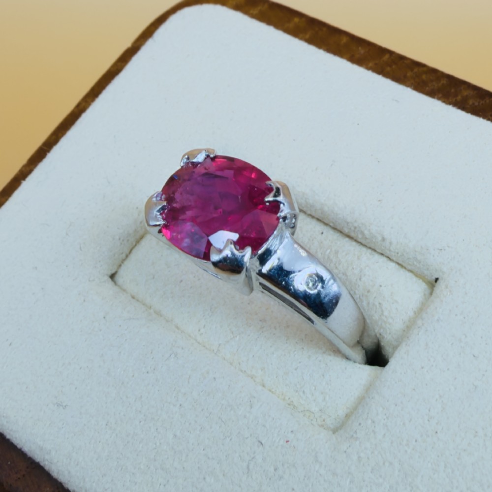 An 18ct white gold ring featuring a prominent oval cut red ruby secured by four corner claws, accented with small diamonds on the shoulders. View III