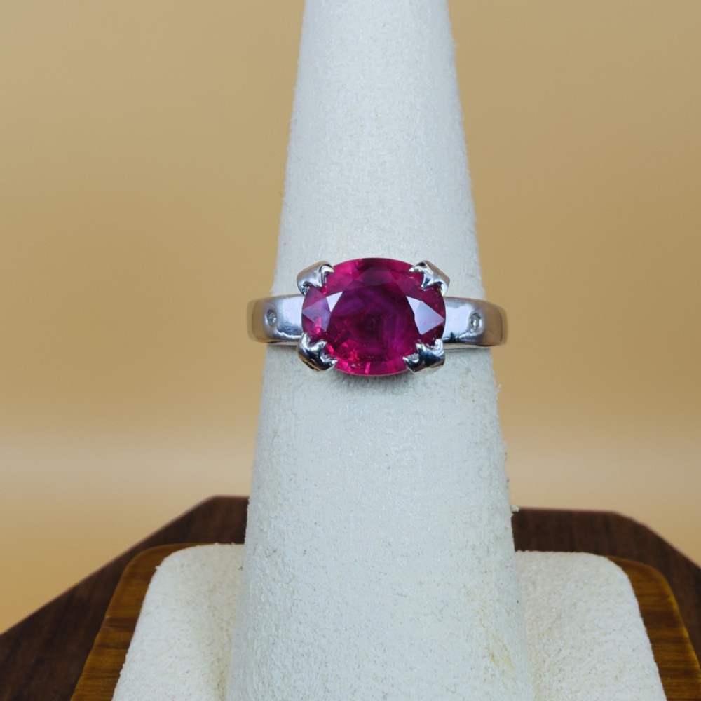 An 18ct white gold ring featuring a prominent oval cut red ruby secured by four corner claws, accented with small diamonds on the shoulders. View VI