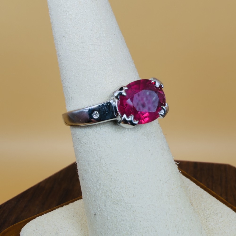 An 18ct white gold ring featuring a prominent oval cut red ruby secured by four corner claws, accented with small diamonds on the shoulders. View VIII