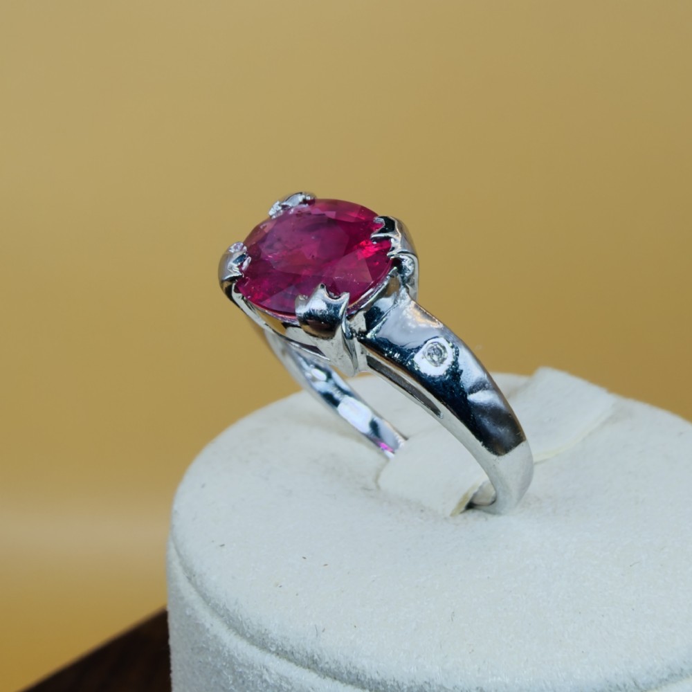 An 18ct white gold ring featuring a prominent oval cut red ruby secured by four corner claws, accented with small diamonds on the shoulders. View XI