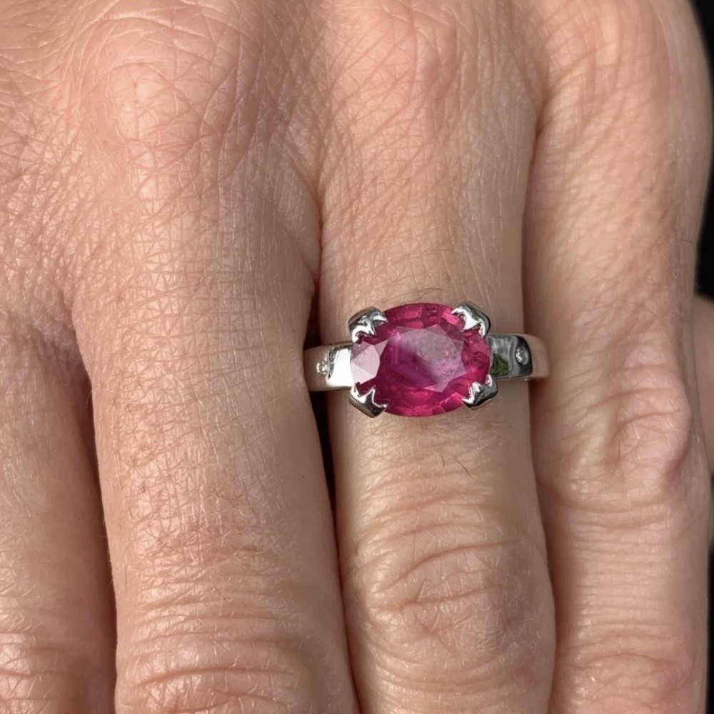An 18ct white gold ring featuring a prominent oval cut red ruby secured by four corner claws, accented with small diamonds on the shoulders. View XII