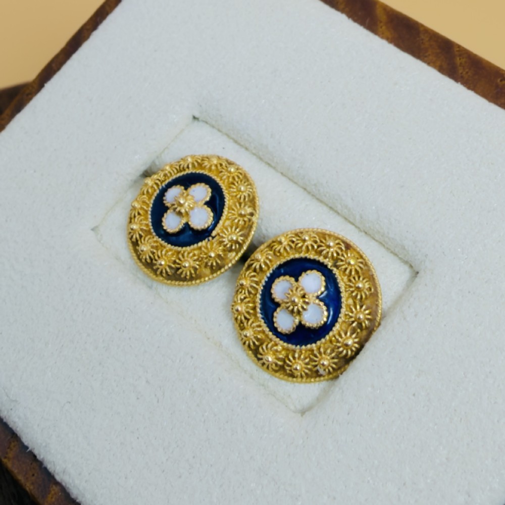 A pair of circular 19.2ct yellow gold stud earrings featuring a central white four-petalled flower against a dark blue enamel ground, surrounded by textured gold detail. View II