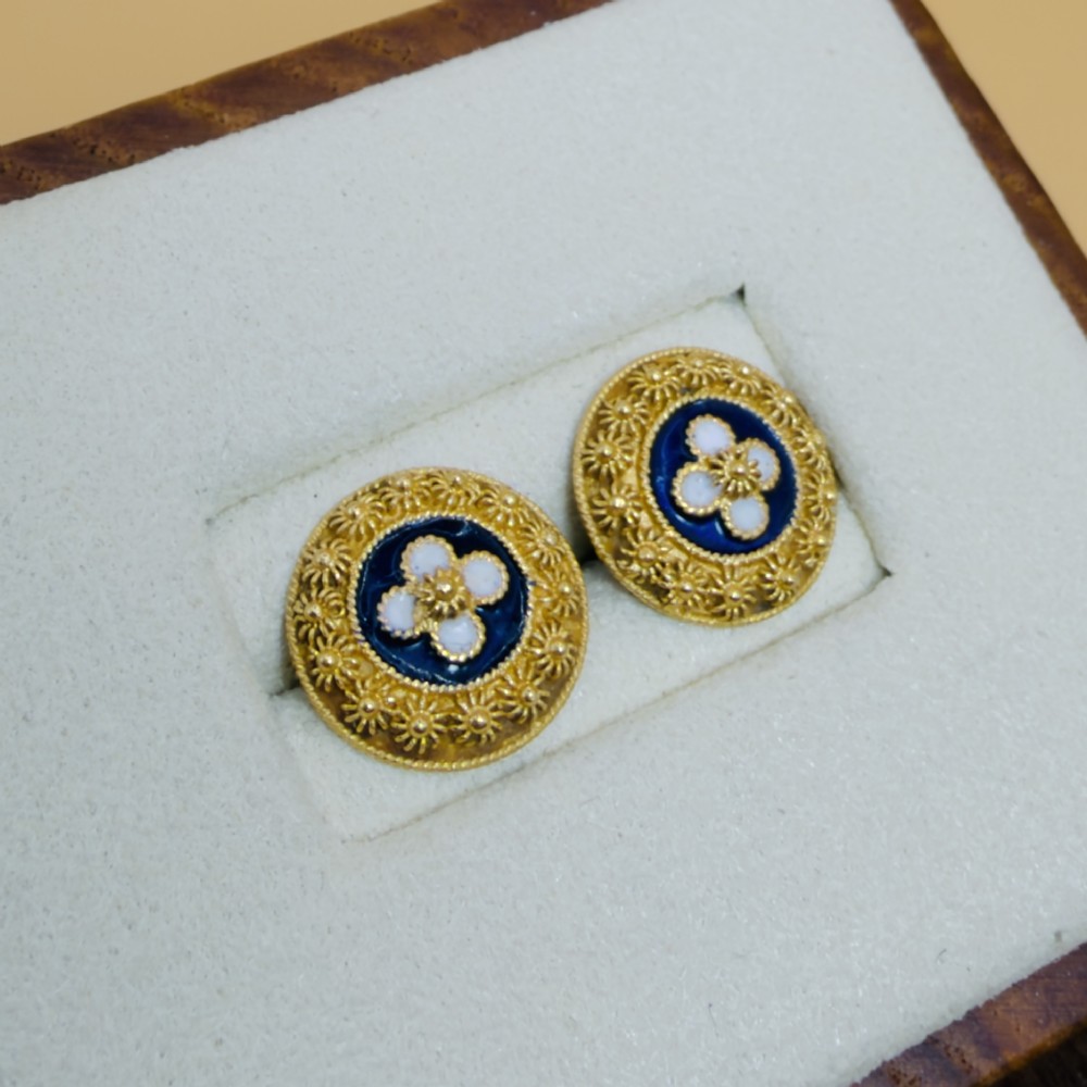A pair of circular 19.2ct yellow gold stud earrings featuring a central white four-petalled flower against a dark blue enamel ground, surrounded by textured gold detail. View III