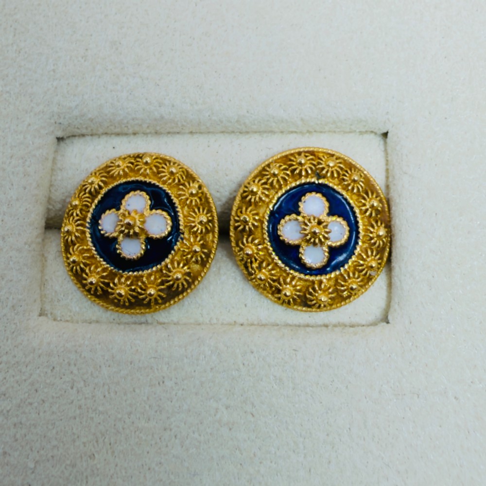 A pair of circular 19.2ct yellow gold stud earrings featuring a central white four-petalled flower against a dark blue enamel ground, surrounded by textured gold detail. View IV