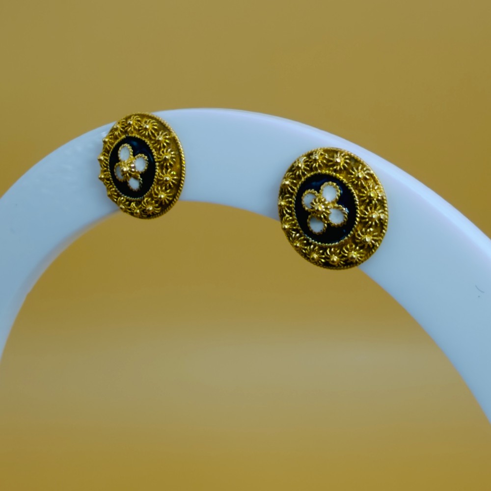 A pair of circular 19.2ct yellow gold stud earrings featuring a central white four-petalled flower against a dark blue enamel ground, surrounded by textured gold detail. View VI
