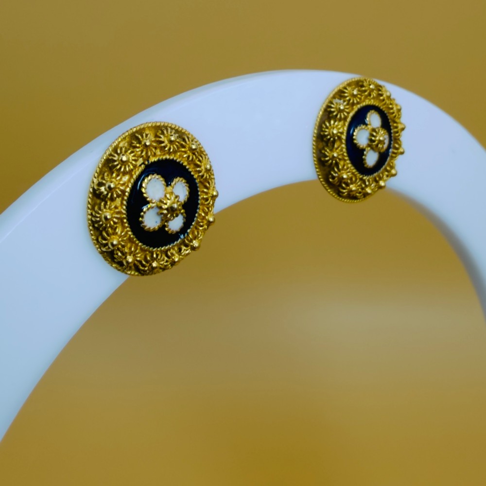A pair of circular 19.2ct yellow gold stud earrings featuring a central white four-petalled flower against a dark blue enamel ground, surrounded by textured gold detail. View VII