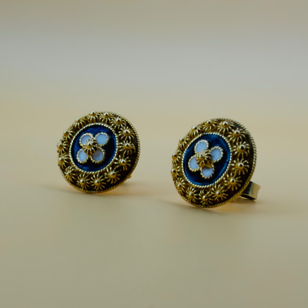 A pair of circular 19.2ct yellow gold stud earrings featuring a central white four-petalled flower against a dark blue enamel ground, surrounded by textured gold detail. View VIII