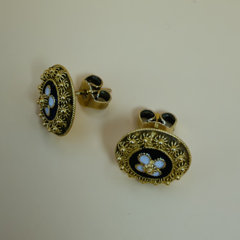 A pair of circular 19.2ct yellow gold stud earrings featuring a central white four-petalled flower against a dark blue enamel ground, surrounded by textured gold detail. View IX
