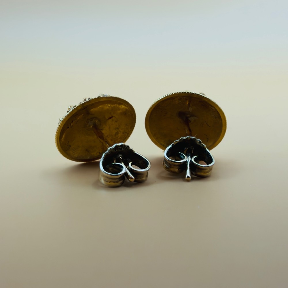 A pair of circular 19.2ct yellow gold stud earrings featuring a central white four-petalled flower against a dark blue enamel ground, surrounded by textured gold detail. View X