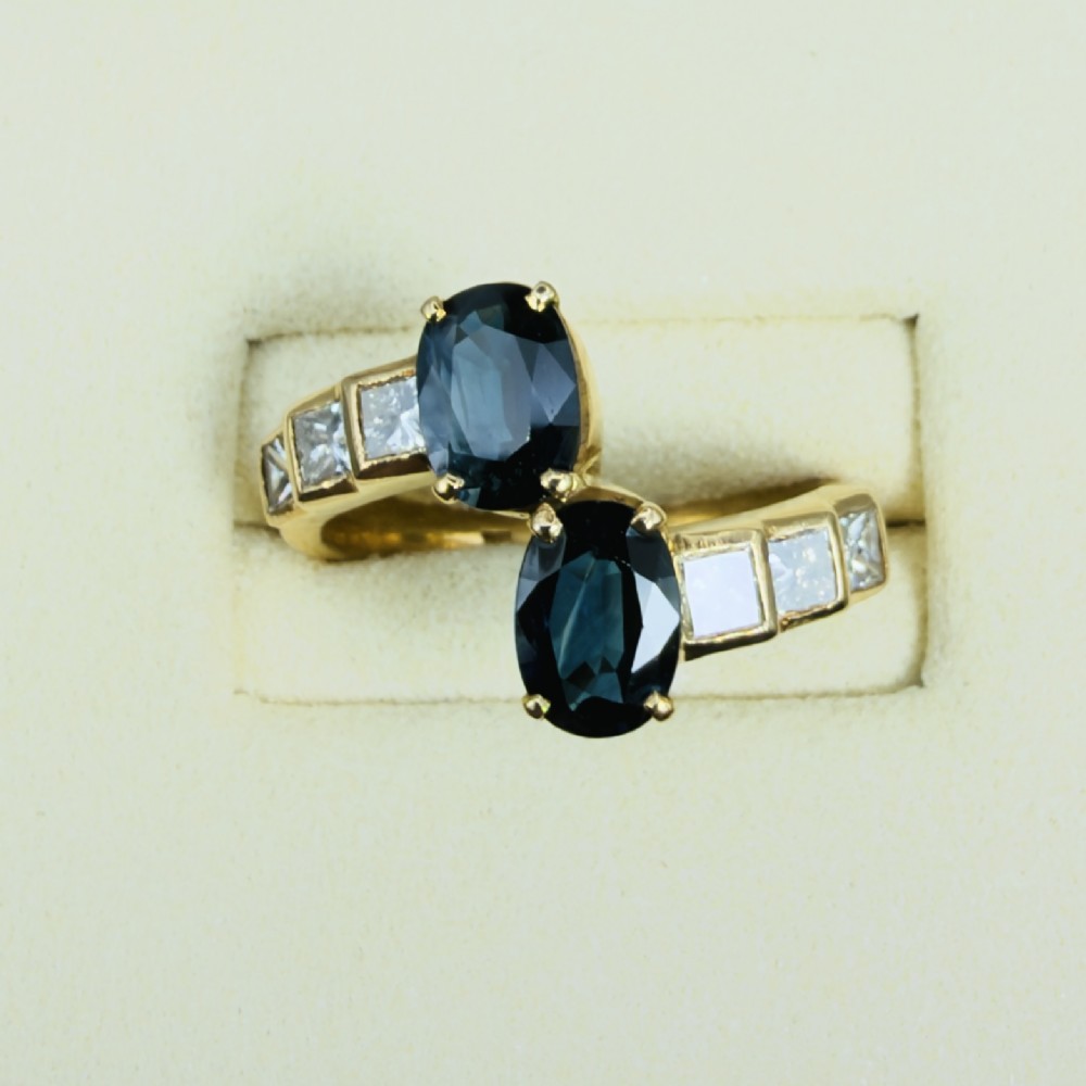 An 18ct yellow gold Art Deco ring featuring two oval-cut blue sapphires in a crossover setting with stepped shoulders set with princess-cut diamonds. View II