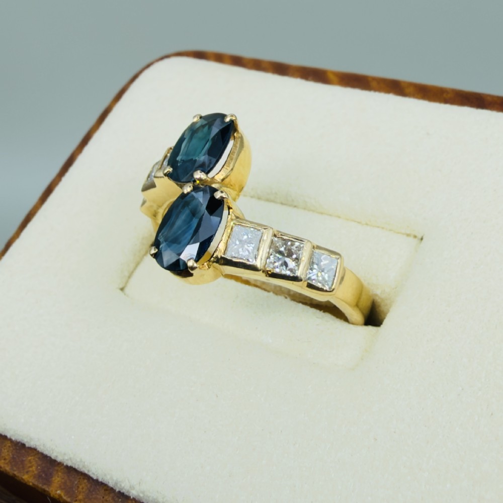 An 18ct yellow gold Art Deco ring featuring two oval-cut blue sapphires in a crossover setting with stepped shoulders set with princess-cut diamonds. View III