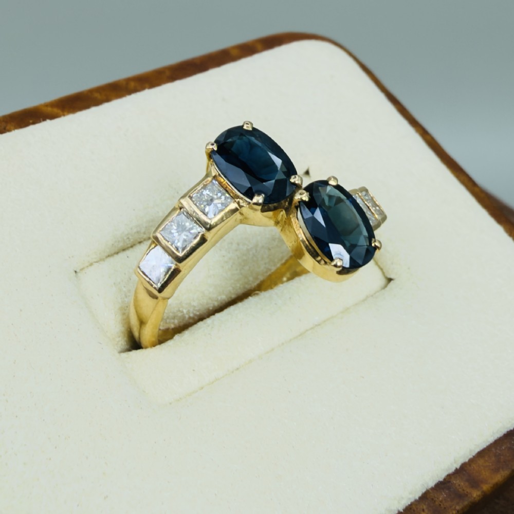 An 18ct yellow gold Art Deco ring featuring two oval-cut blue sapphires in a crossover setting with stepped shoulders set with princess-cut diamonds. View IV