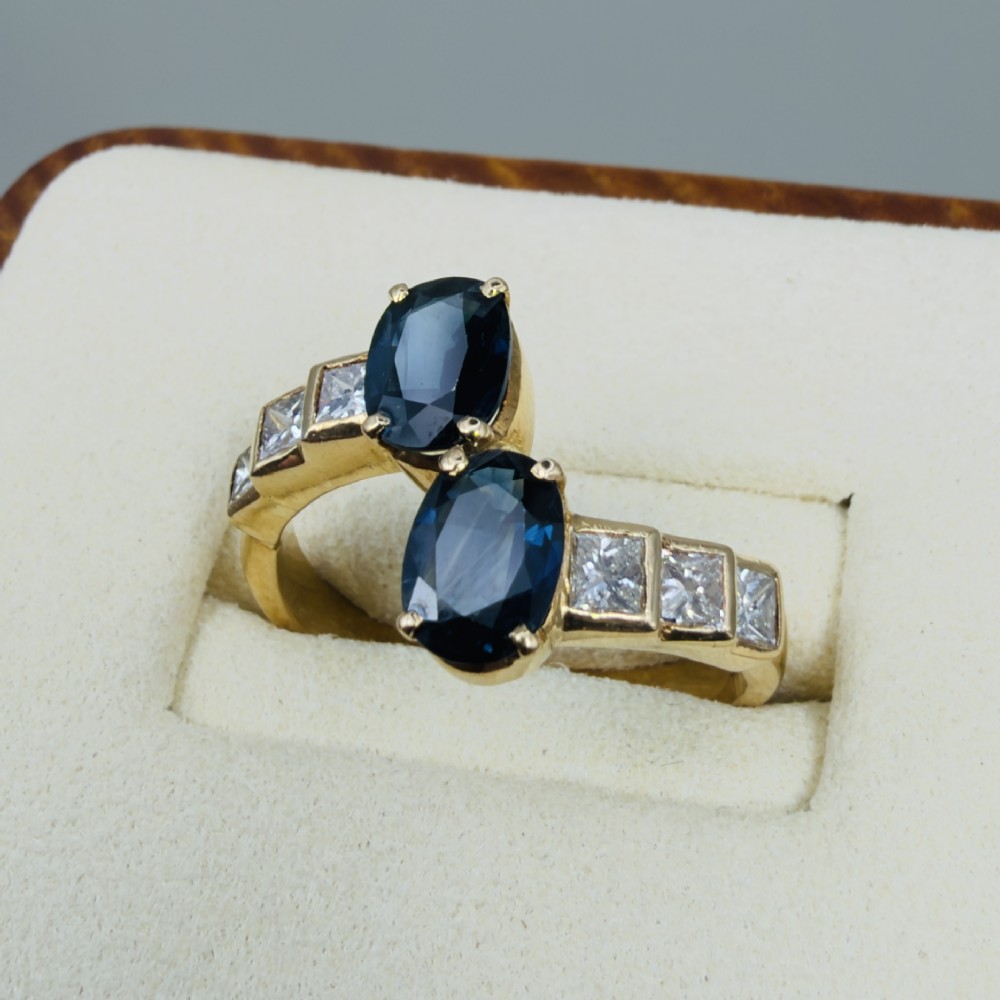 An 18ct yellow gold Art Deco ring featuring two oval-cut blue sapphires in a crossover setting with stepped shoulders set with princess-cut diamonds. View V
