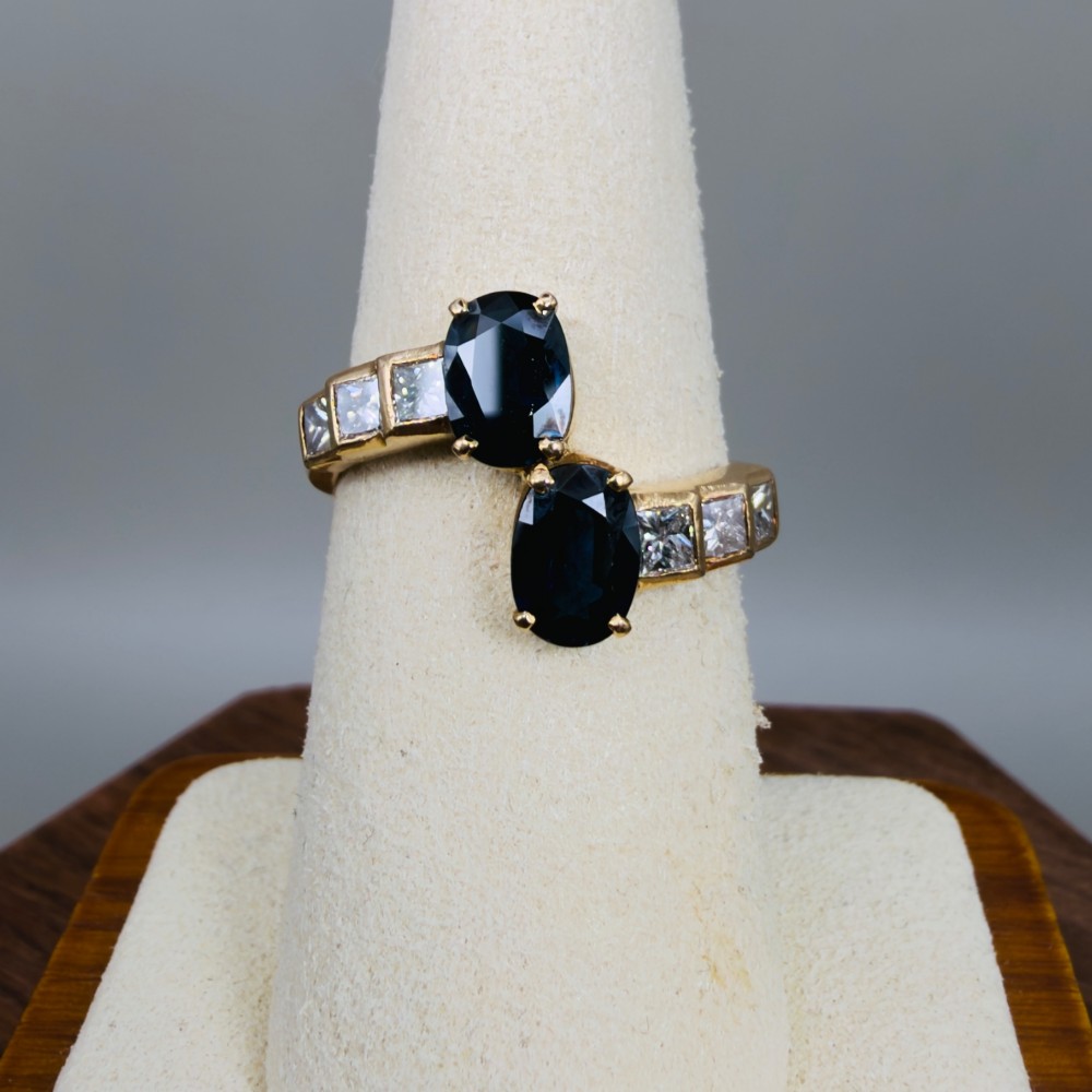 An 18ct yellow gold Art Deco ring featuring two oval-cut blue sapphires in a crossover setting with stepped shoulders set with princess-cut diamonds. View VI