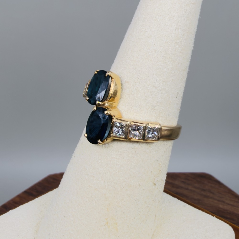 An 18ct yellow gold Art Deco ring featuring two oval-cut blue sapphires in a crossover setting with stepped shoulders set with princess-cut diamonds. View VII
