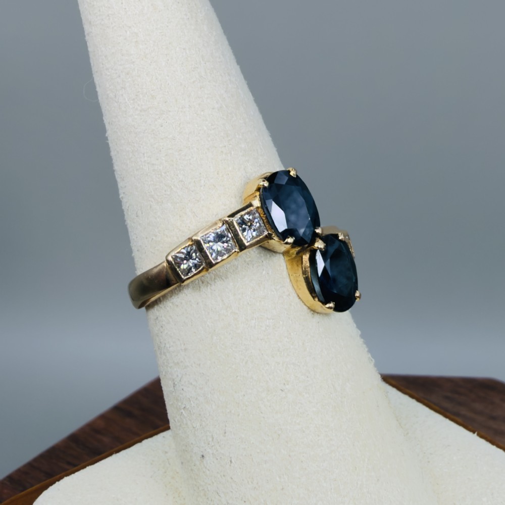 An 18ct yellow gold Art Deco ring featuring two oval-cut blue sapphires in a crossover setting with stepped shoulders set with princess-cut diamonds. View VIII