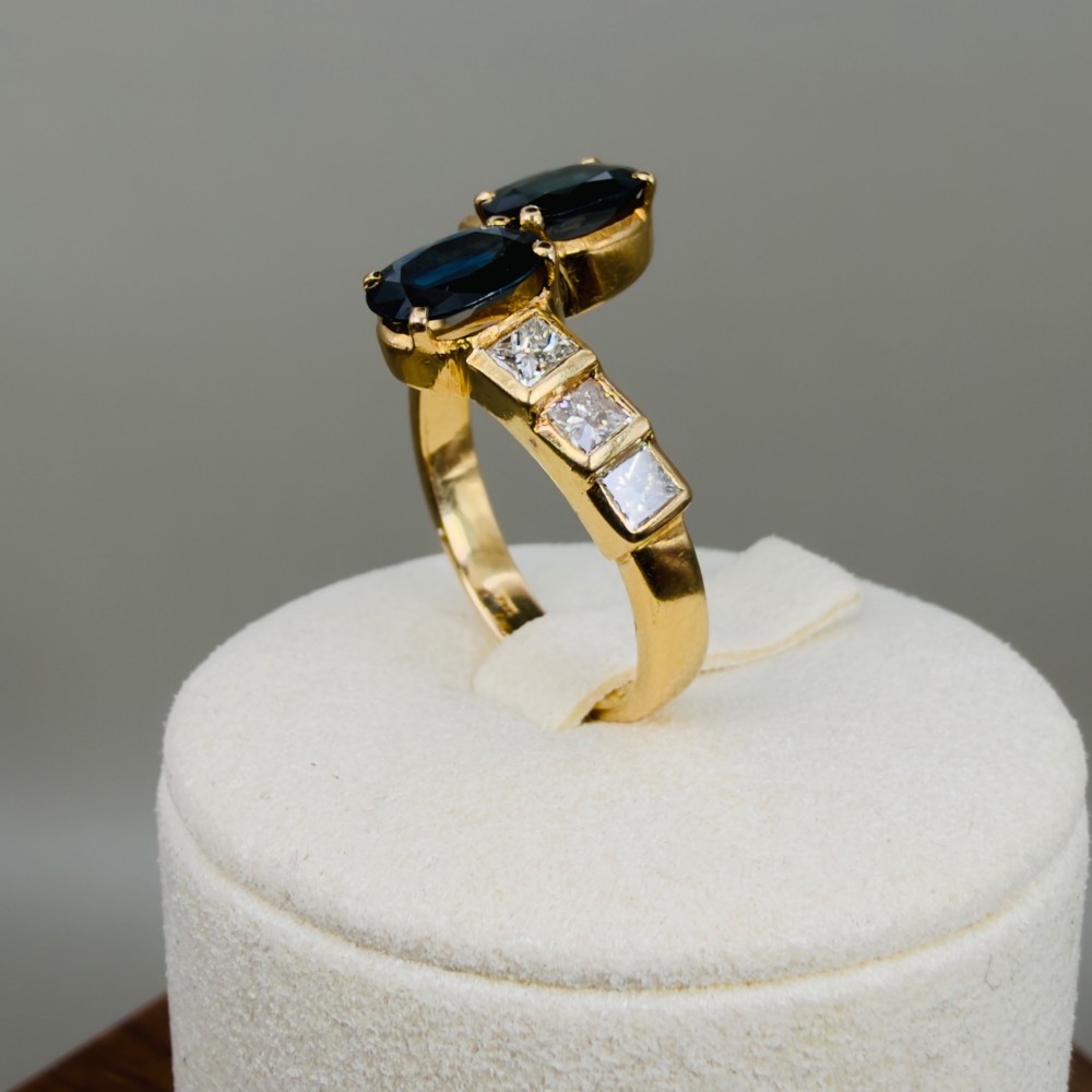 An 18ct yellow gold Art Deco ring featuring two oval-cut blue sapphires in a crossover setting with stepped shoulders set with princess-cut diamonds. View IX