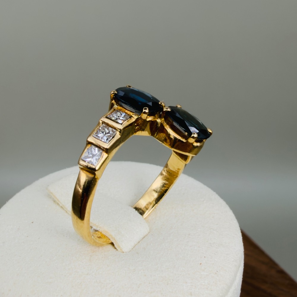 An 18ct yellow gold Art Deco ring featuring two oval-cut blue sapphires in a crossover setting with stepped shoulders set with princess-cut diamonds. View X