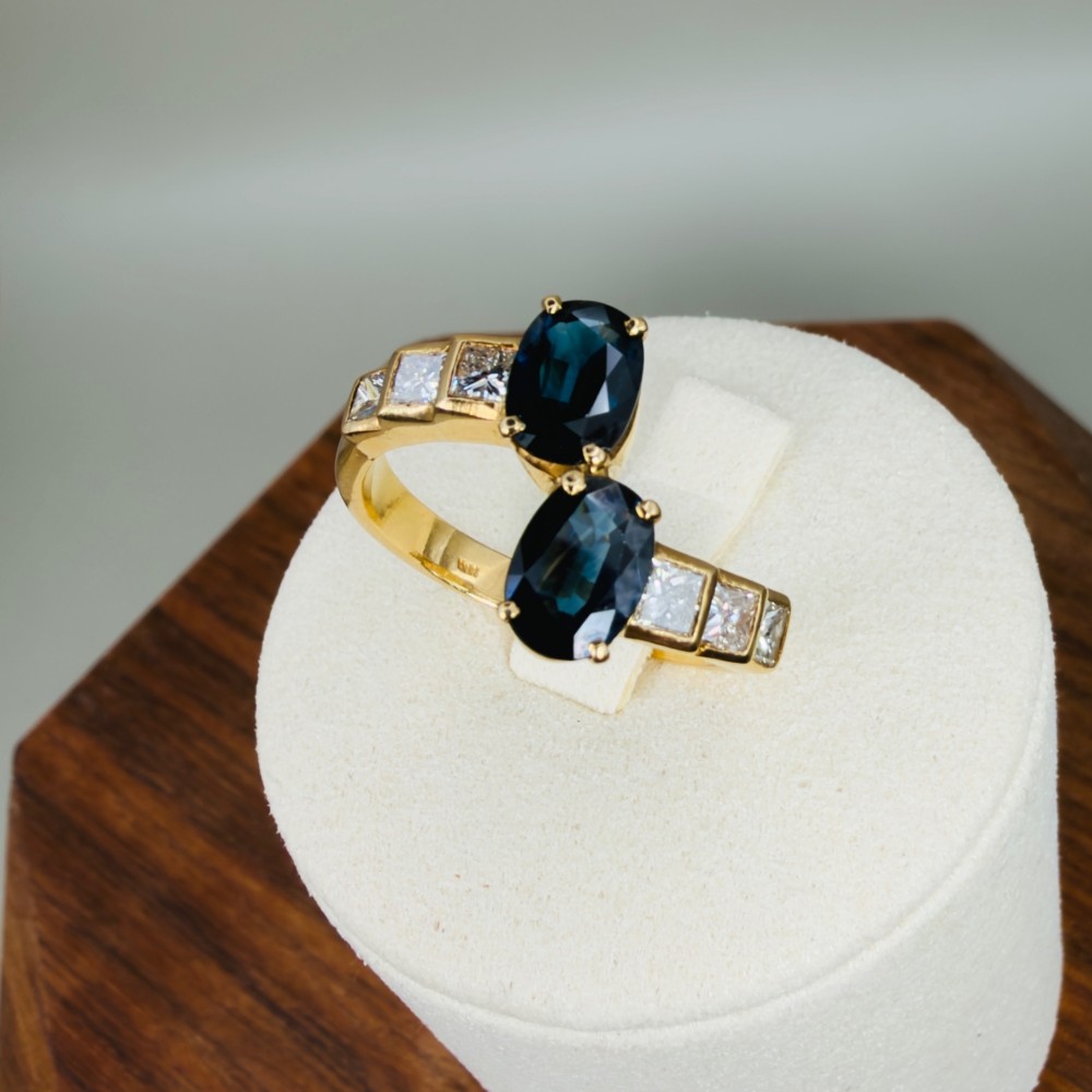An 18ct yellow gold Art Deco ring featuring two oval-cut blue sapphires in a crossover setting with stepped shoulders set with princess-cut diamonds. View XI