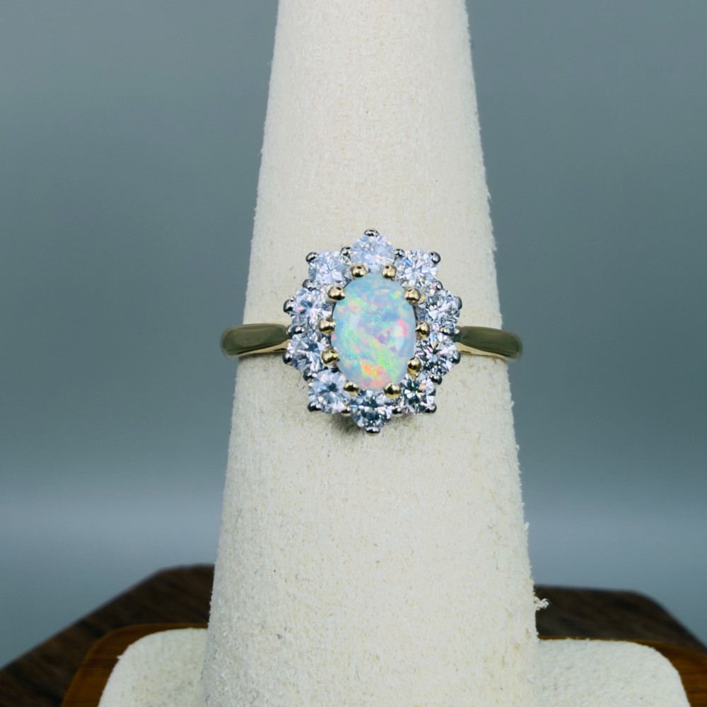An 18ct yellow gold ring featuring an oval cabochon opal surrounded by a halo of brilliant-cut diamonds. View VI