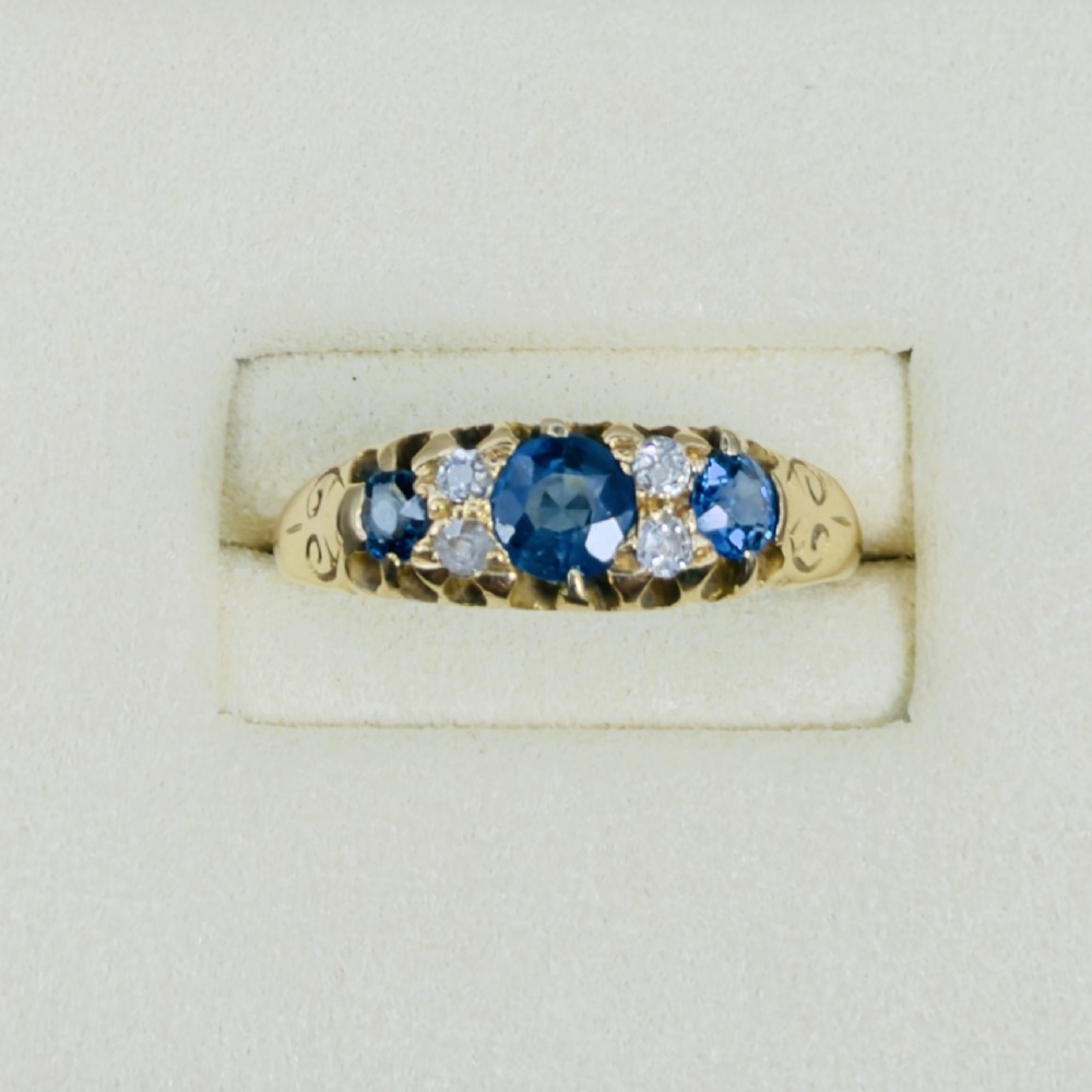 An 18ct yellow gold Edwardian ring set with three oval blue sapphires and four diamond accents featuring carved scroll shoulders. View II