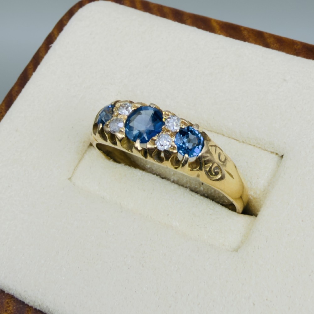 An 18ct yellow gold Edwardian ring set with three oval blue sapphires and four diamond accents featuring carved scroll shoulders. View III