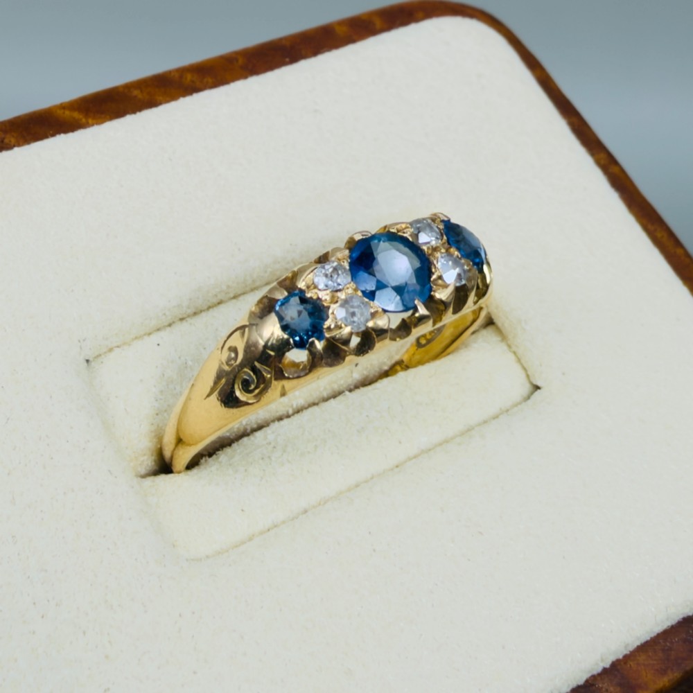An 18ct yellow gold Edwardian ring set with three oval blue sapphires and four diamond accents featuring carved scroll shoulders. View IV