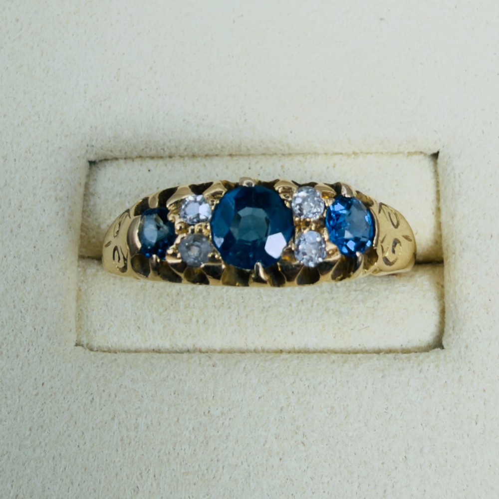 An 18ct yellow gold Edwardian ring set with three oval blue sapphires and four diamond accents featuring carved scroll shoulders. View V