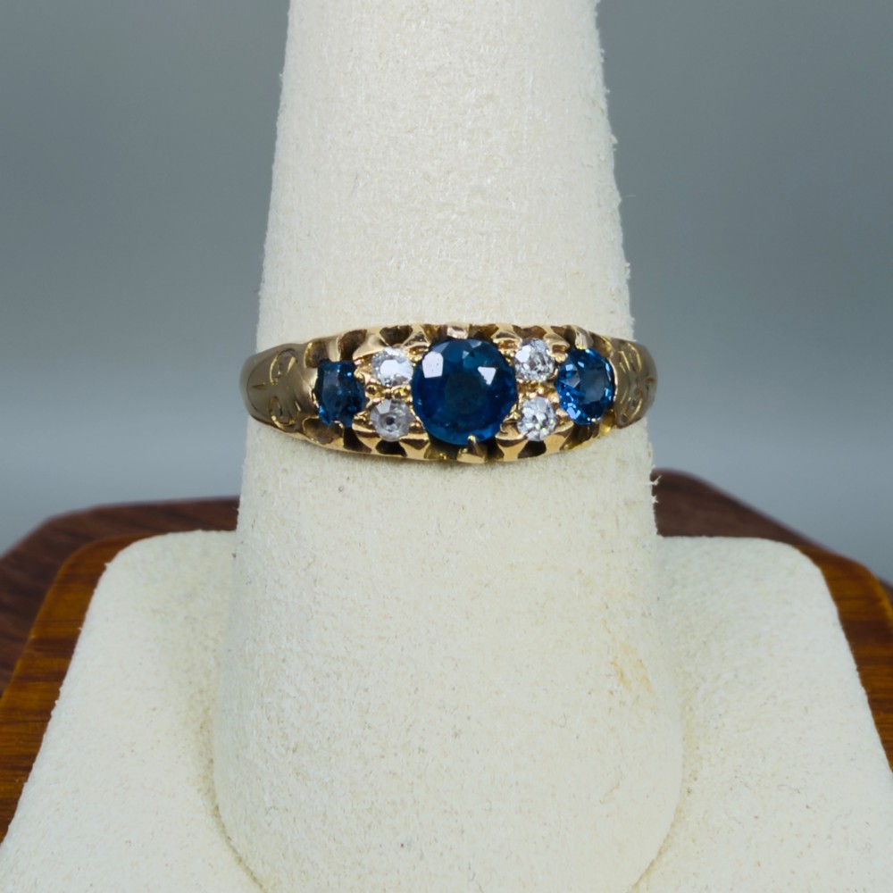 An 18ct yellow gold Edwardian ring set with three oval blue sapphires and four diamond accents featuring carved scroll shoulders. View VI
