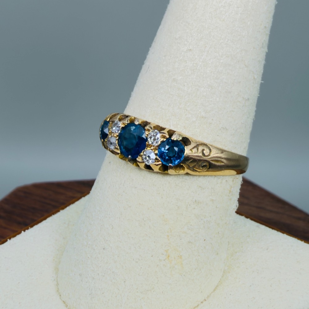An 18ct yellow gold Edwardian ring set with three oval blue sapphires and four diamond accents featuring carved scroll shoulders. View VII