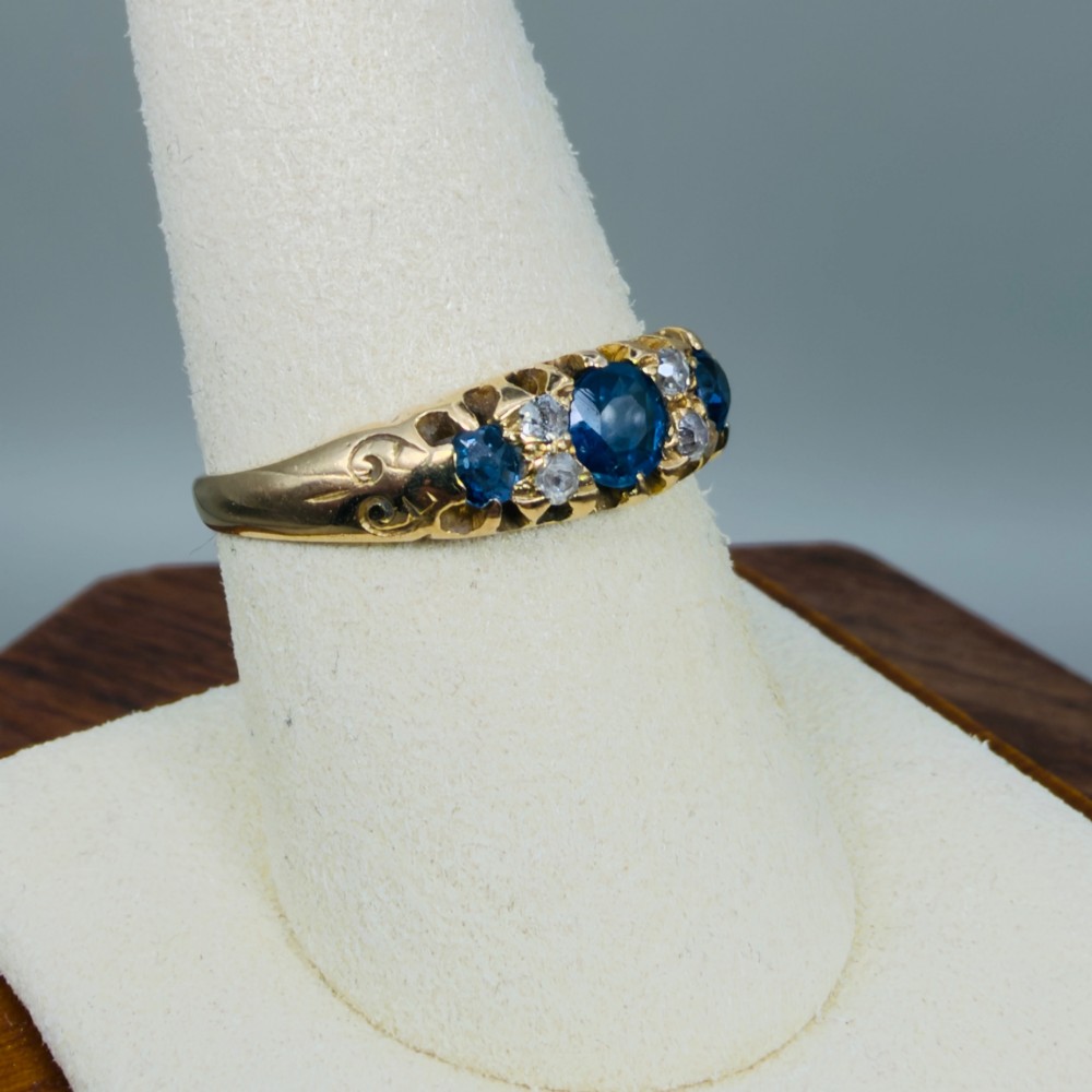 An 18ct yellow gold Edwardian ring set with three oval blue sapphires and four diamond accents featuring carved scroll shoulders. View VIII