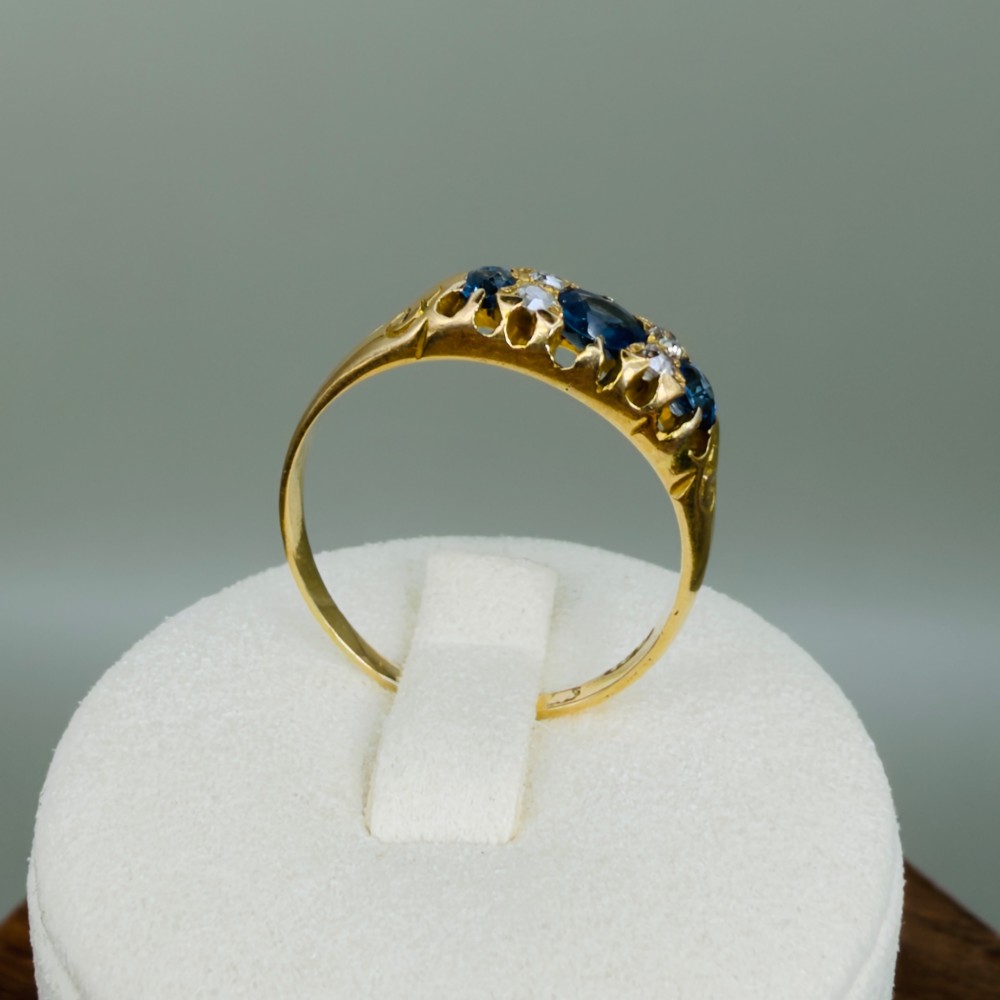 An 18ct yellow gold Edwardian ring set with three oval blue sapphires and four diamond accents featuring carved scroll shoulders. View IX