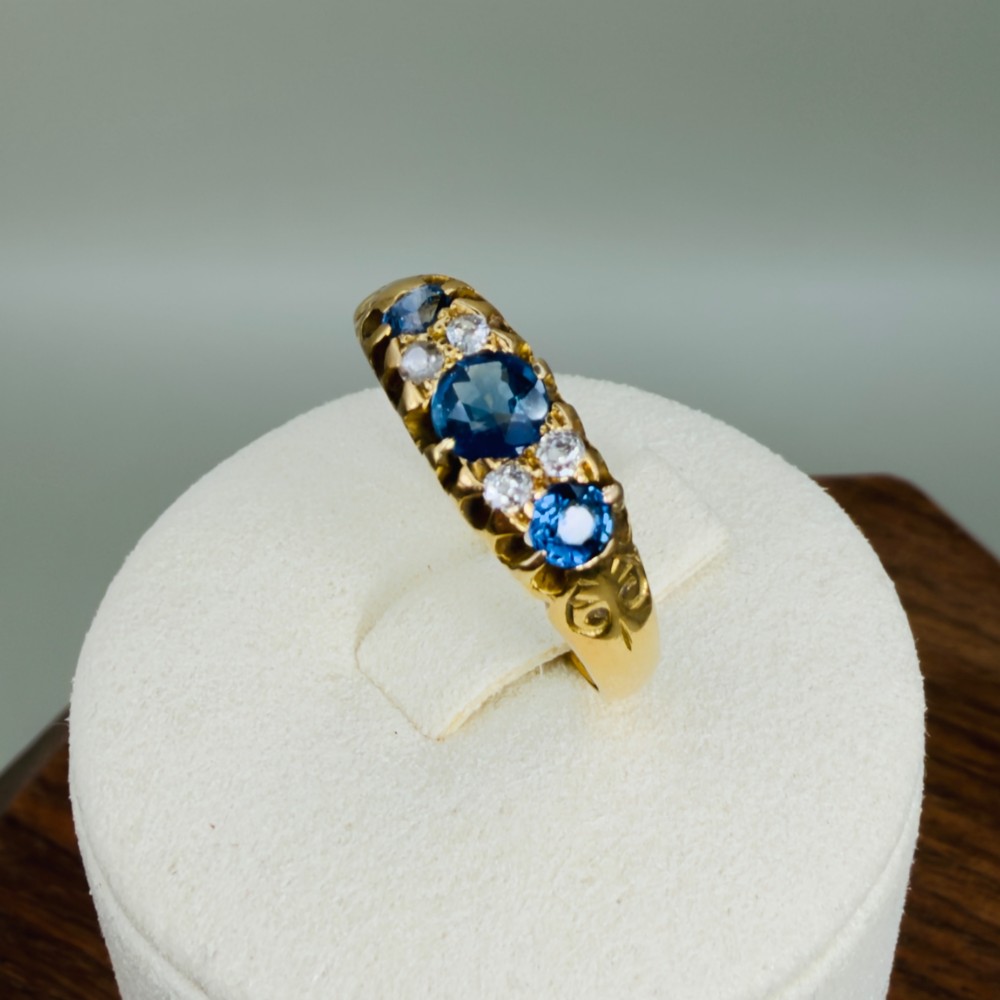 An 18ct yellow gold Edwardian ring set with three oval blue sapphires and four diamond accents featuring carved scroll shoulders. View X