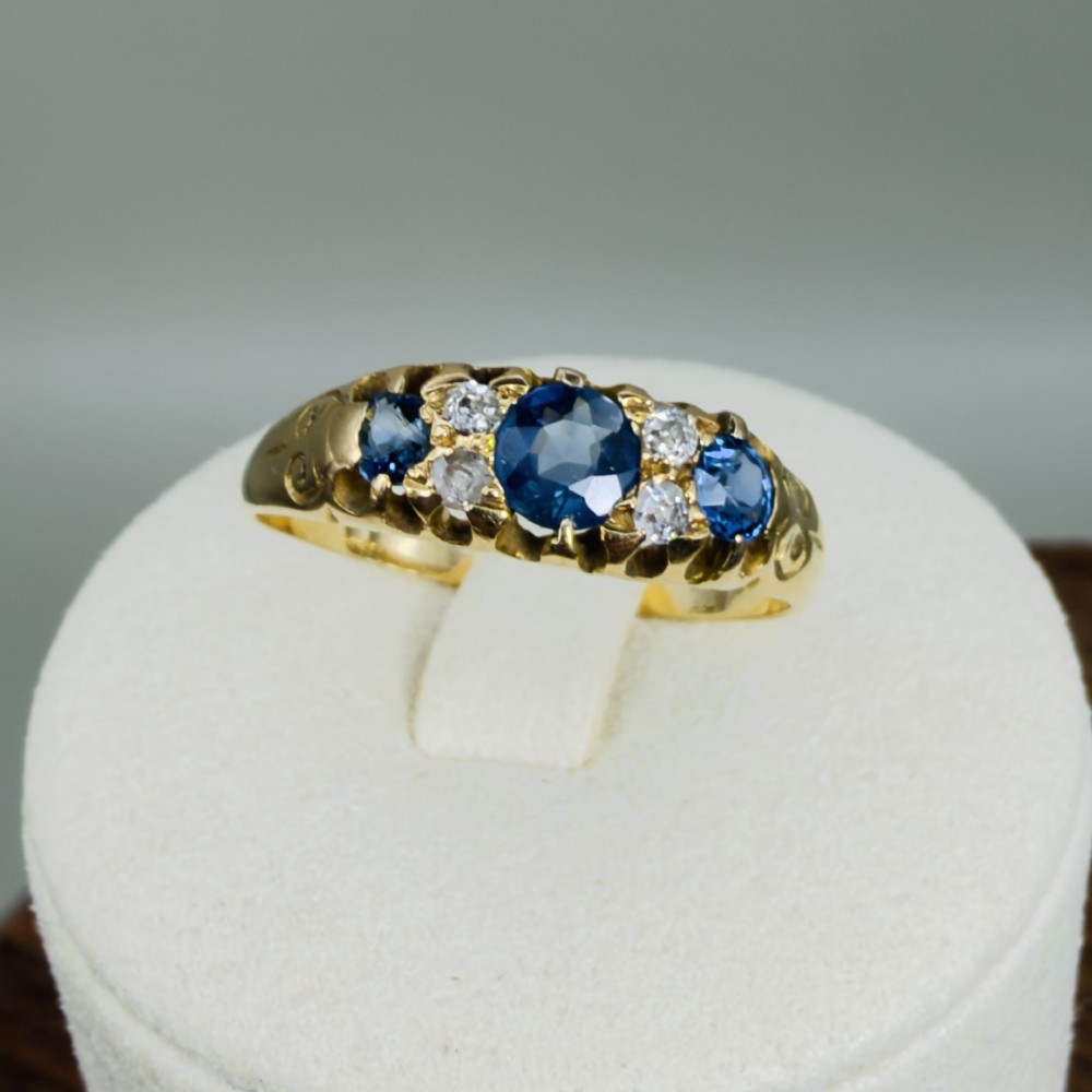 An 18ct yellow gold Edwardian ring set with three oval blue sapphires and four diamond accents featuring carved scroll shoulders.