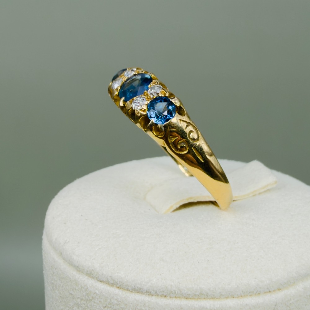 An 18ct yellow gold Edwardian ring set with three oval blue sapphires and four diamond accents featuring carved scroll shoulders. View XI